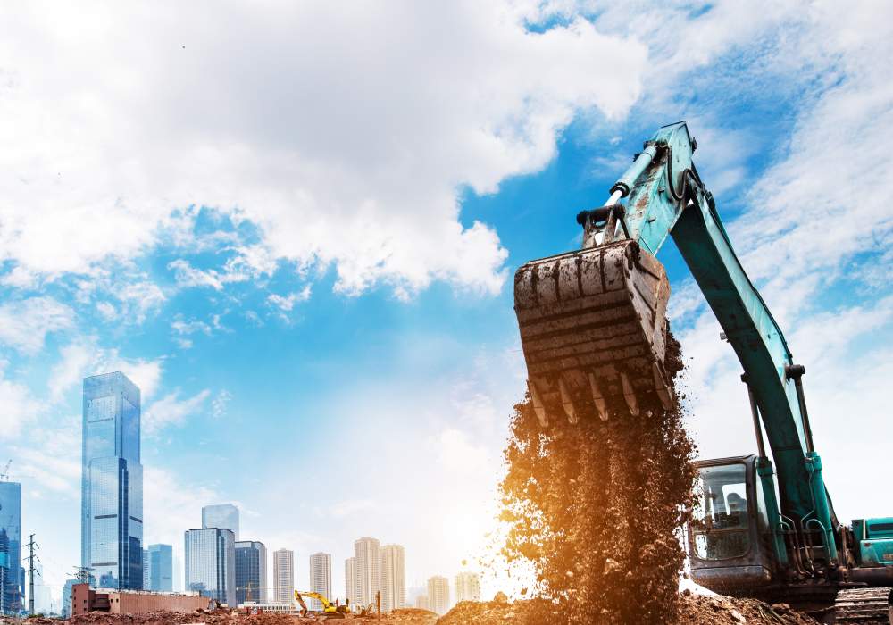 Excavator working on construction site near city buildings under blue sky